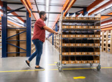 A young warehouse employee pushing a picking cart past aisles of merchandise in a fulfillment center.